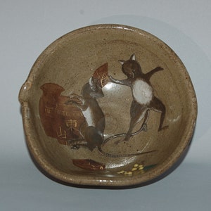 May include: A tan ceramic bowl featuring a playful illustration of two mice. One mouse is brown and white, holding a fan, the other is grey and white, near a brown vase with Japanese characters. The bowl has a small spout.