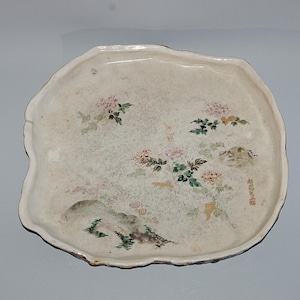 May include: A vintage, off-white ceramic serving platter with an irregular edge. The platter is decorated with hand-painted floral and landscape motifs in shades of pink, green, and yellow. The design includes flowers, rocks, and a small inscription.