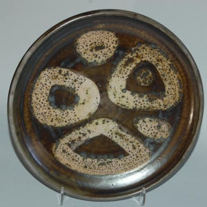 May include: A ceramic plate with a brown and cream speckled design. The plate has a dark brown centre with cream-coloured shapes and a dark brown rim. The plate is on a clear stand.