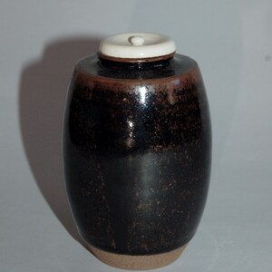 Könnte beinhalten: Eine schwarze Keramikvase mit einem weißen Deckel. Die Vase hat eine gesprenkelte braune Glasur und eine abgerundete Form.