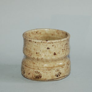 May include: A cylindrical ceramic cup with a beige speckled glaze and a smooth finish. The base features a horizontal band decorated with a repeating embossed floral pattern. The cup has a slightly indented middle section and a wide, open rim.