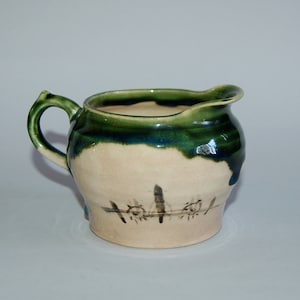 May include: Handmade ceramic creamer with a green and blue glazed rim and handle. The off-white body features a dark, linear design. A unique, rustic piece for serving.