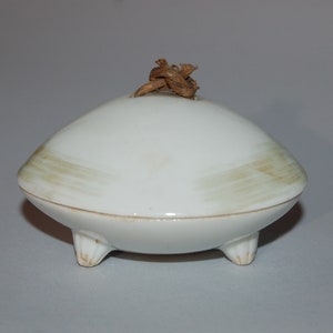 Op de afbeelding: Een kleine, witte, koepelvormige keramische pot met deksel en drie korte, stompe pootjes. Het deksel heeft een lichtbruin, horizontaal penseelstreekpatroon en is in het midden voorzien van een kleine, geknoopte handgreep van natuurlijke vezels.