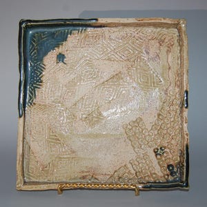 Op de afbeelding: Een vierkant keramisch dienblad met een reliëf geometrisch patroon en een blauwe glazuur langs de randen. Het dienblad heeft een getextureerd oppervlak en een witte glazuur.