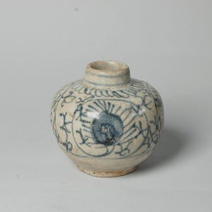 May include: A small, round, antique ceramic vase with a short neck. The off-white vase features blue floral patterns and a central blue design. The vase is likely hand-painted and has a rustic, aged appearance.