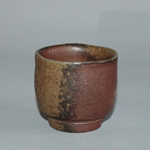 May include: A handmade ceramic cup with a rustic appearance. The cup features a two-tone design, with a textured brown exterior and a reddish-brown interior. The cup has a rounded shape and a slightly uneven rim.