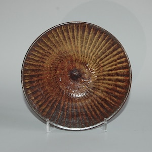 Puede incluir: Un plato redondo de cerámica con un patrón radial texturizado en tonos marrones y beige. El plato tiene un borde marrón oscuro y una depresión central más oscura. Se exhibe sobre un soporte transparente.