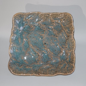 Op de afbeelding: Vierkante keramische schaal met een getextureerd oppervlak en onregelmatige randen. De schaal heeft een gevlekte blauwgroene en bruine glazuur. Het handgemaakte aardewerk heeft een rustieke, organische uitstraling.