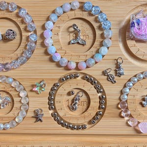 May include: A wooden bead board with circular channels containing various jewelry components including clear crackle glass beads, pastel stone beads, white pearls, silver metallic beads, and assorted charms like a whale tail, turtle, and starfish.