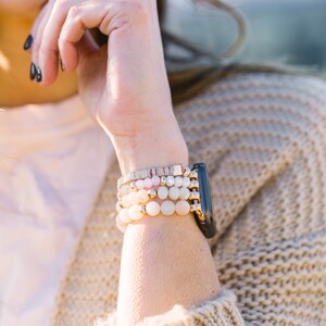 May include: A stack of beaded bracelets in shades of pink, white, and gold, paired with a black smartwatch. The bracelets feature various bead sizes and textures, creating a layered look. The watch has a gold band.