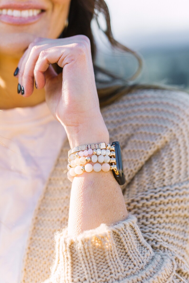 May include: A stack of beaded bracelets in shades of pink, white, and gold, paired with a black smartwatch. The bracelets feature various bead sizes and textures, creating a layered look. The watch has a gold band.