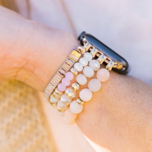 May include: A collection of beaded bracelets in shades of white, pink, and gold, stacked on a wrist next to a black smartwatch. The bracelets feature various bead sizes and textures, creating a layered look.