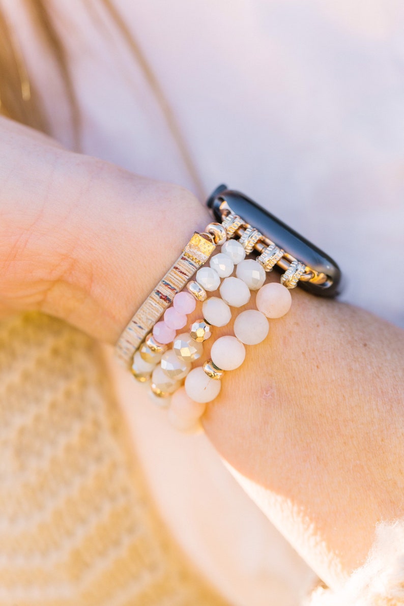 May include: A collection of beaded bracelets in shades of white, pink, and gold, stacked on a wrist next to a black smartwatch. The bracelets feature various bead sizes and textures, creating a layered look.