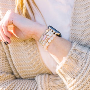 May include: A wrist adorned with a black smartwatch and multiple beaded bracelets in shades of white, pink, and gold. The bracelets feature various textures and bead sizes. The person is wearing a beige knit cardigan.