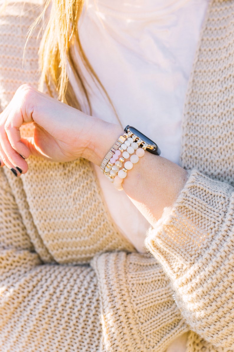 May include: A wrist adorned with a black smartwatch and multiple beaded bracelets in shades of white, pink, and gold. The bracelets feature various textures and bead sizes. The person is wearing a beige knit cardigan.