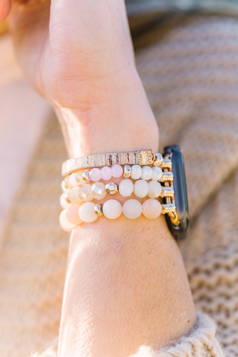 May include: A stack of beaded bracelets in shades of pink, cream, and gold, worn with a black smartwatch. The bracelets feature various bead sizes and textures, including round and faceted beads.
