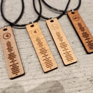 Op de afbeelding: Vier houten kettingen met zwarte koorden. Elke hanger is een rechthoekig stuk hout met een geluidgolfpatroon en een klein symbool bovenaan. De symbolen zijn een hart, een noot, een cirkel en een hart.