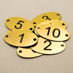 May include: A group of oval-shaped, gold-coloured metal tags with black numbers. The numbers on the tags are 1, 2, 3, 5, and 10.