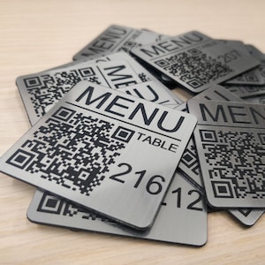 Puede incluir: Un montón de letreros de número de mesa de metal plateado con códigos QR negros y la palabra "MENU" impresa en ellos. Los números de mesa son 216, 217 y 207.