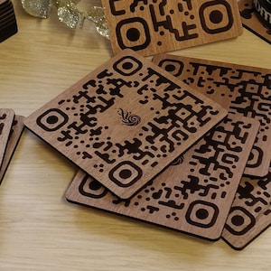 Può includere: Un set di sei sottobicchieri in legno con un design a codice QR in bianco e nero. I sottobicchieri sono quadrati e hanno angoli arrotondati. I sottobicchieri sono impilati con il sottobicchiere superiore rivolto verso la fotocamera. I sottobicchieri sono su una superficie in legno.