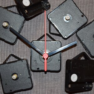 May include: A set of six black clock mechanisms with a red second hand. The clock mechanisms have a black metal frame and a white plastic gear. The mechanisms are arranged in a circle on a gray surface.