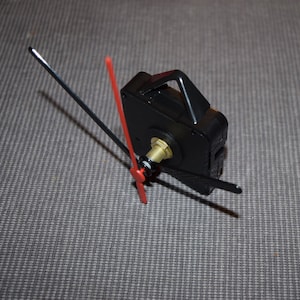 May include: A black clock mechanism with a gold winding stem and two clock hands, one red and one black, attached.