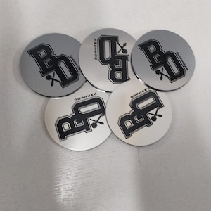 May include: Five silver round coasters with a black logo of the letters "BB" with a crossed spoon and fork design. The logo is in a bold, block font.