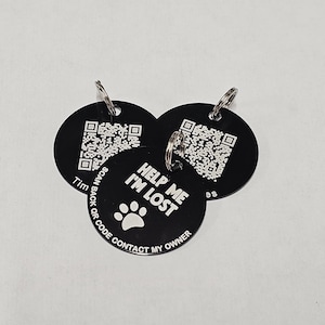 May include: Three black round pet ID tags with silver key rings. The tags have a QR code and the text "Help Me I'm Lost" with a paw print. The text on the bottom of the tag reads "Scan Back QR Code Contact My Owner".