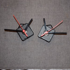 May include: Two black clock mechanisms with red hands. The hands are attached to a square base with a small hole in the center.