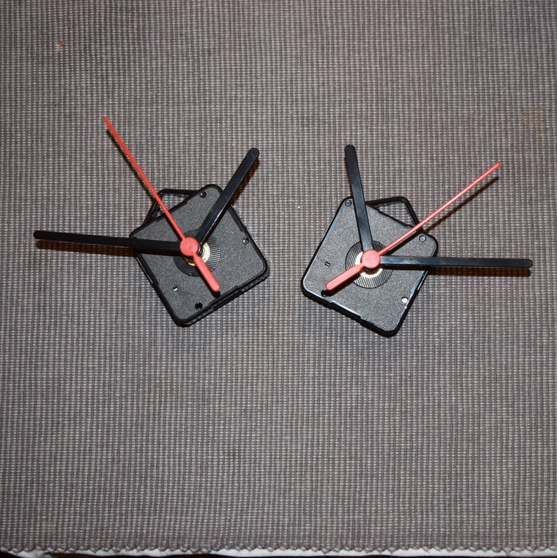 May include: Two black clock mechanisms with red hands. The hands are attached to a square base with a small hole in the center.
