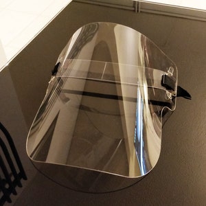 May include: A clear plastic face shield with a black elastic band. The shield is curved and has a wide field of view.