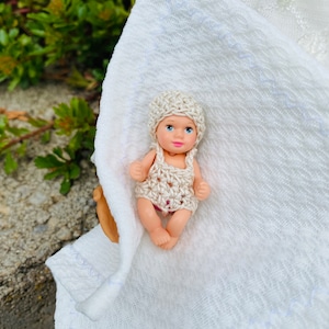 May include: A small doll wearing a crocheted outfit and hat. The doll has blue eyes and pink lips. The doll is lying on a white cloth with a textured pattern. The background is blurred with green foliage.