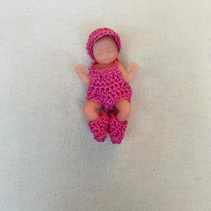 May include: A small doll wearing a handmade, bright pink crochet outfit, including a hat, romper, and booties. The doll has a realistic skin tone and is lying down with its eyes closed, suggesting sleep.