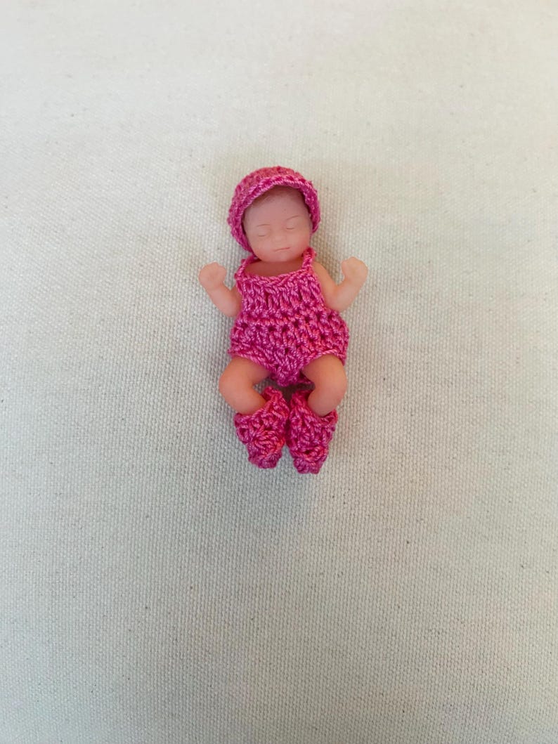 May include: A small doll wearing a handmade, bright pink crochet outfit, including a hat, romper, and booties. The doll has a realistic skin tone and is lying down with its eyes closed, suggesting sleep.