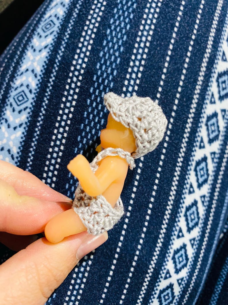 May include: A small, doll-like figurine of a baby wearing a crocheted hat and diaper cover. The hat and diaper cover are light gray. The baby figurine is a light orange color.