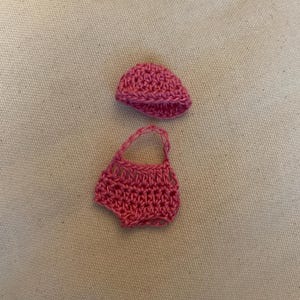 May include: A handmade, pink crochet outfit for a baby, including a small hat and a pair of shorts. The outfit is made of a textured yarn and is displayed on a neutral background.