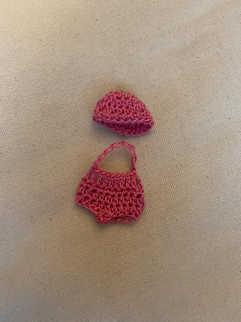 May include: A handmade, pink crochet outfit for a baby, including a small hat and a pair of shorts. The outfit is made of a textured yarn and is displayed on a neutral background.