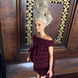 May include: A doll with blonde hair styled in a high bun, wearing a burgundy off-the-shoulder crochet dress. The doll is standing in front of a dark wooden background.
