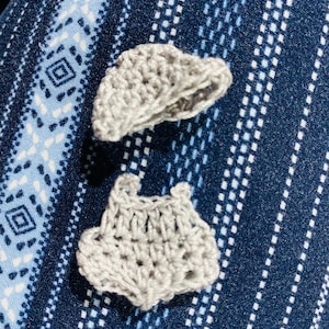 May include: Two small, handmade, light gray crochet items are displayed against a blue patterned fabric background. One item appears to be a small hat, and the other resembles a pair of shorts. The crochet work is visible, showcasing the texture and craftsmanship.