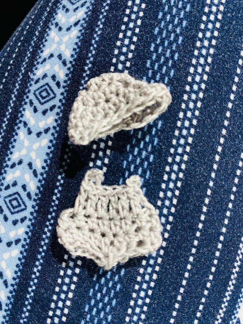 May include: Two small, handmade, light gray crochet items are displayed against a blue patterned fabric background. One item appears to be a small hat, and the other resembles a pair of shorts. The crochet work is visible, showcasing the texture and craftsmanship.