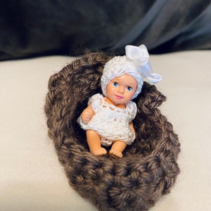 May include: A small white crocheted doll wearing a white crocheted hat with a white bow, lying in a brown crocheted basket.