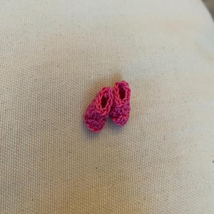 May include: A pair of tiny, handmade, hot pink crochet baby shoes. The shoes are small, with a rounded toe and a simple, open design. The crochet work is tight and even, giving the shoes a neat appearance. The shoes are set against a neutral background.
