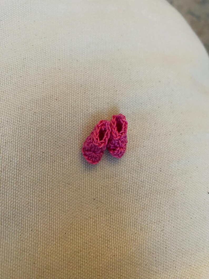 May include: A pair of tiny, handmade, hot pink crochet baby shoes. The shoes are small, with a rounded toe and a simple, open design. The crochet work is tight and even, giving the shoes a neat appearance. The shoes are set against a neutral background.