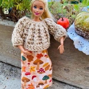 Op de afbeelding: Een pop draagt een beige kabelgebreide trui en een rok met een herfstthema. De rok heeft paddenstoelen, bladeren en andere herfstelementen in bruin-, oranje- en groentinten. De pop draagt ook beige laarzen.