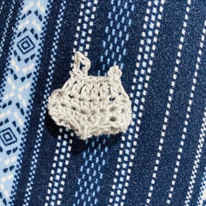 May include: A small, handmade, light gray crochet item, possibly a baby garment, is centered in the image. The crochet work has an open, airy design. The background is a blue fabric with a white geometric pattern.