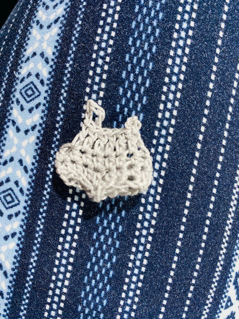 May include: A small, handmade, light gray crochet item, possibly a baby garment, is centered in the image. The crochet work has an open, airy design. The background is a blue fabric with a white geometric pattern.