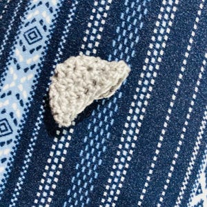 May include: A small, crocheted gray object, possibly a decorative element or applique, rests on a patterned blue fabric. The fabric features a repeating design of white and light blue geometric shapes and lines, creating a textured background.