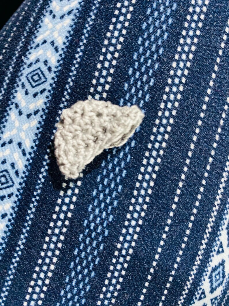 May include: A small, crocheted gray object, possibly a decorative element or applique, rests on a patterned blue fabric. The fabric features a repeating design of white and light blue geometric shapes and lines, creating a textured background.