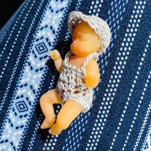 May include: A small, doll-like figurine of a baby wearing a crocheted outfit and hat. The baby is lying down, with its arms and legs slightly extended. The outfit and hat are a light gray color. The baby's skin is a pale peach color.