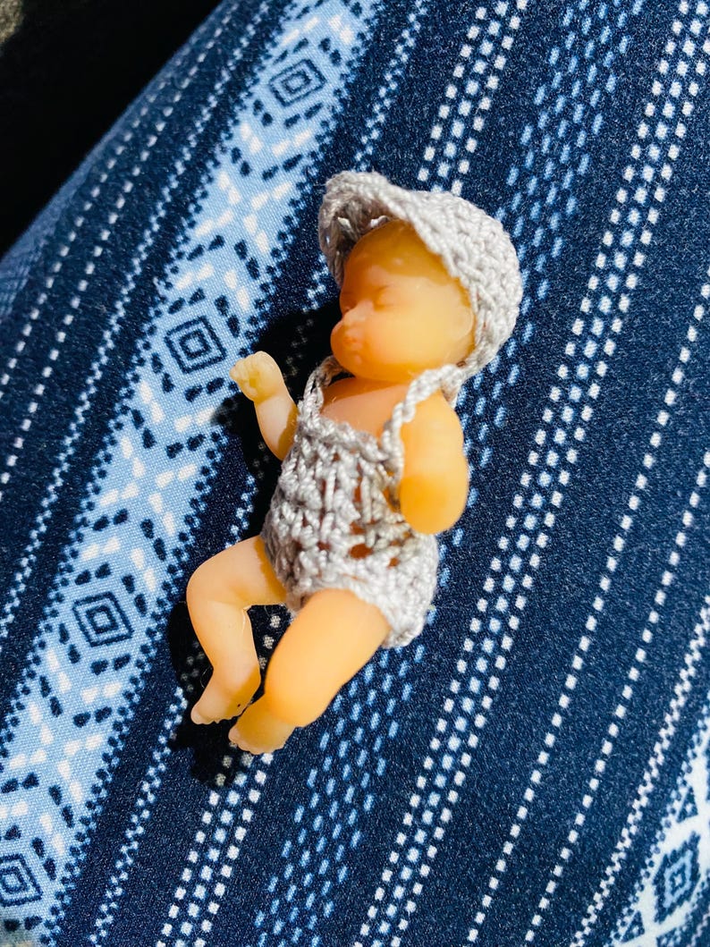 May include: A small, doll-like figurine of a baby wearing a crocheted outfit and hat. The baby is lying down, with its arms and legs slightly extended. The outfit and hat are a light gray color. The baby's skin is a pale peach color.